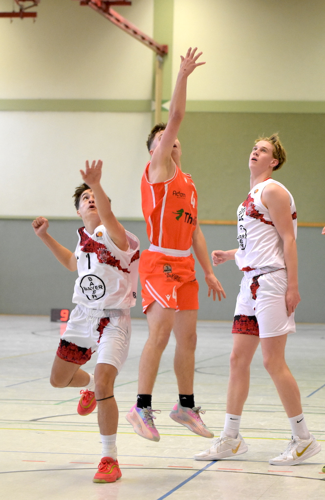 Drei junge Basketballspieler, einer in Orange beim Sprungwurf, konzentrierte Gesichter