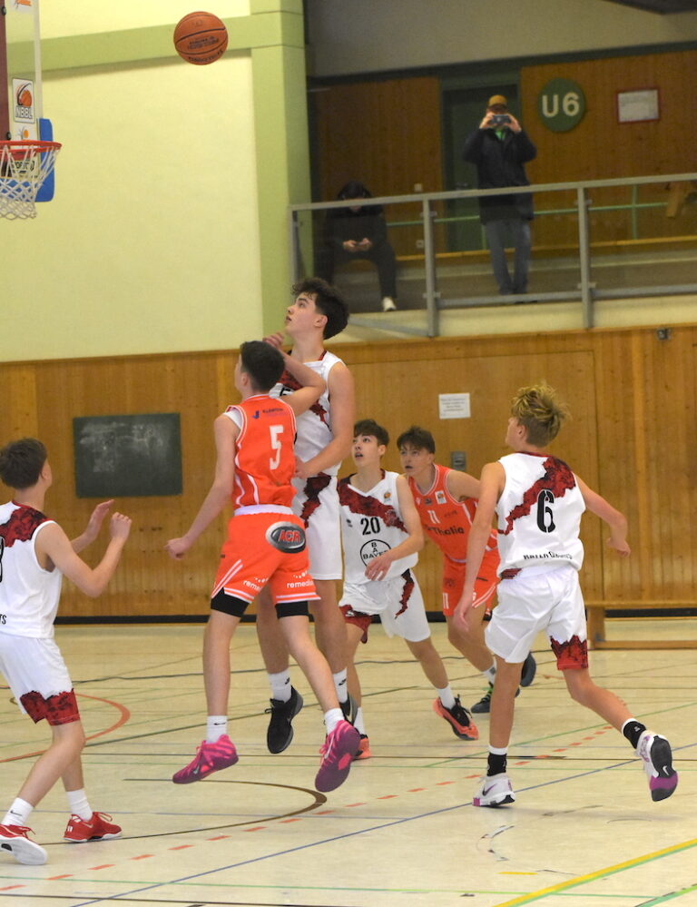 Mehrere Jungen spielen Basketball in einer Turnhalle, einer springt zum Korb