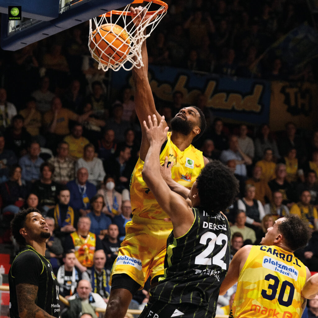 Ein Basketballspieler im gelben Trikot dunkt entschlossen über Verteidiger in Schwarz