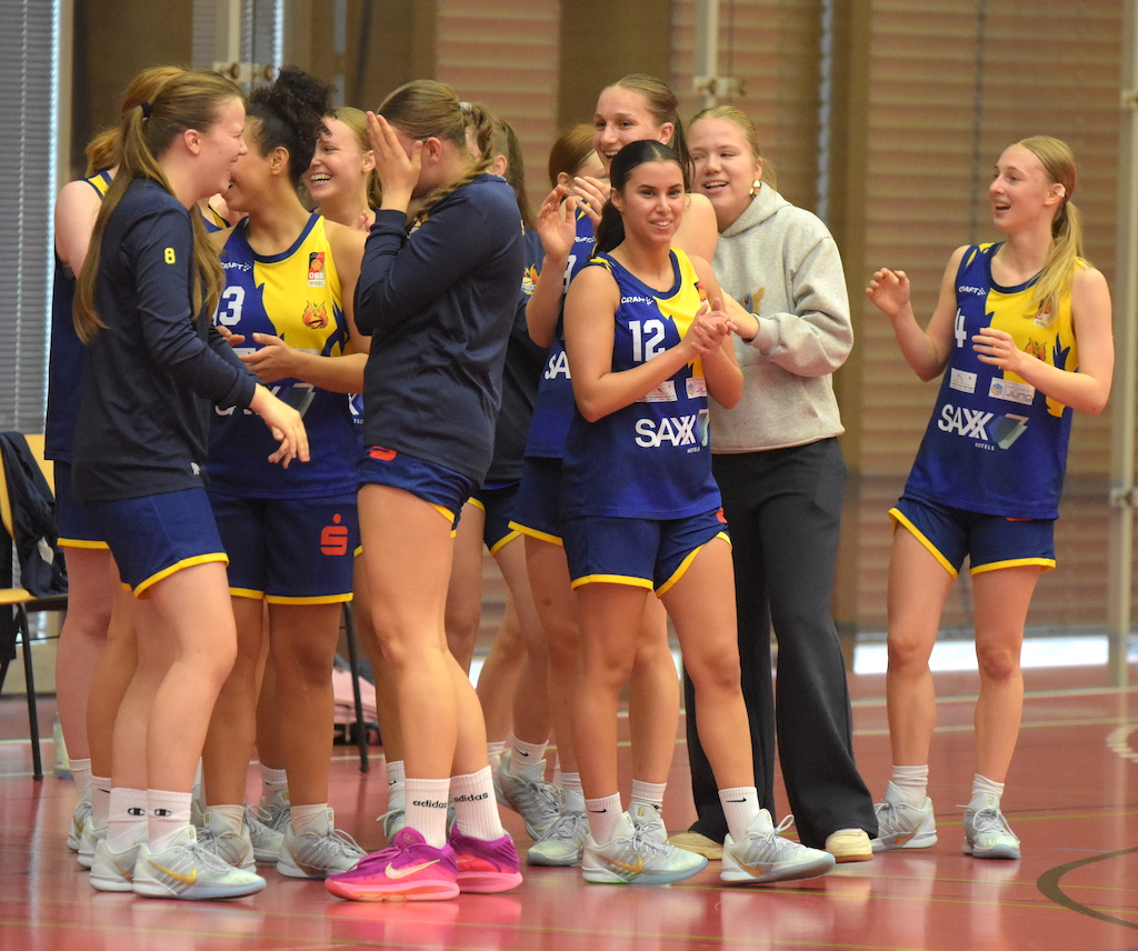 Eine Gruppe junger Basketballspielerinnen lacht zusammen in blauen Trikots auf dem Spielfeld