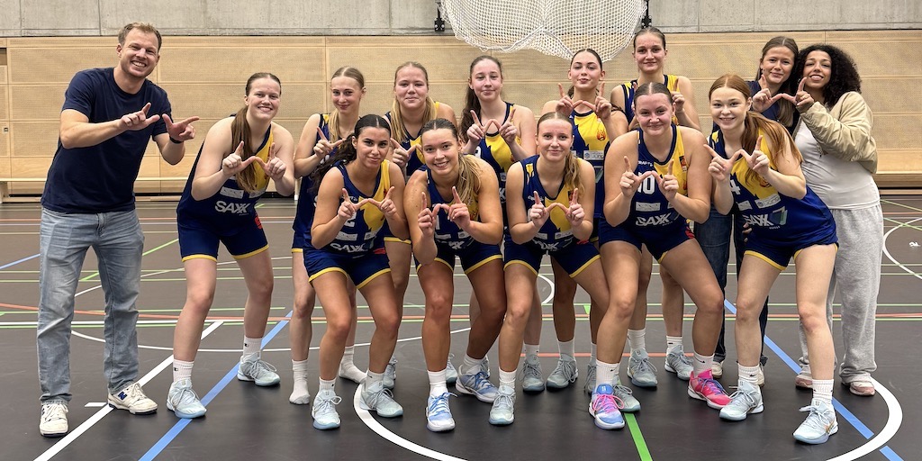 Eine Damen Basketballmannschaft in blauen Trikots posiert lächelnd in Sporthalle
