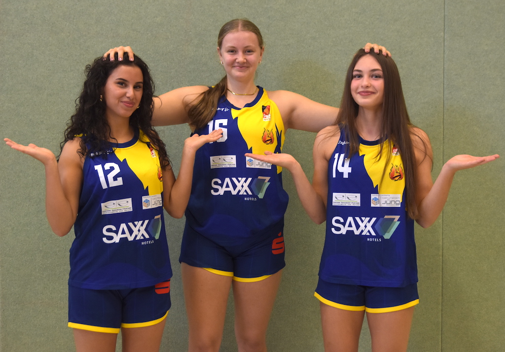 Drei junge Basketballspielerinnen in blaugelben Trikots posieren lächelnd vor grüner Wand