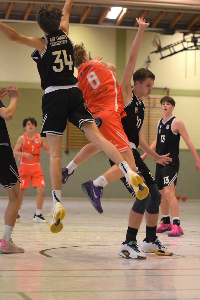 Junger Basketballspieler im orangenen Trikot springt dynamisch zwischen Gegnern in Schwarz