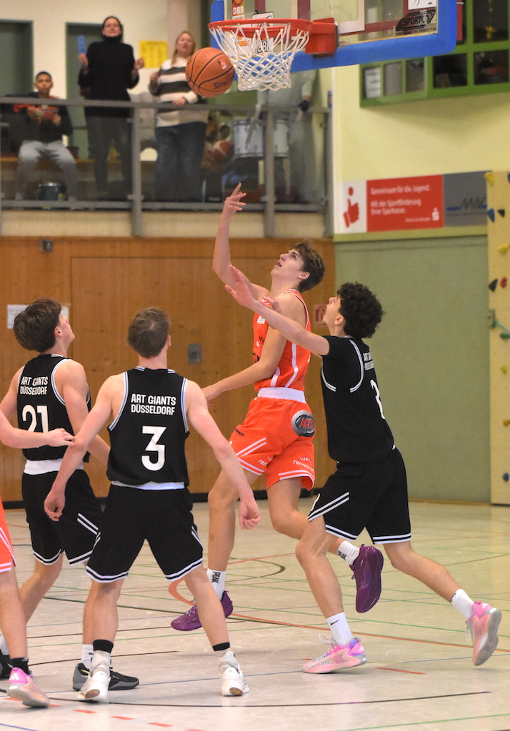 Ein Basketballspieler im roten Trikot springt konzentriert zum Korb, drei Spieler in schwarzen Trikots verteidigen energisch, im Hintergrund jubeln Zuschauer auf einer Empore