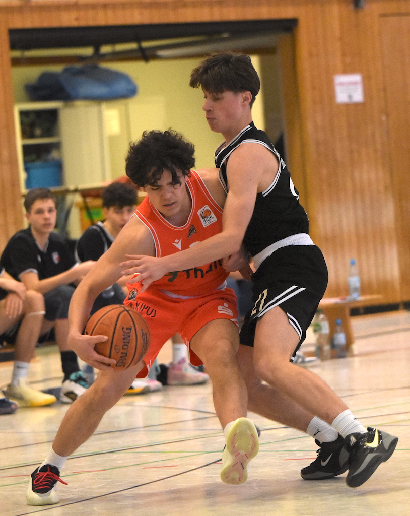 Zwei Jugendliche spielen Basketball in einer Halle, einer in Orange setzt zum Korb an