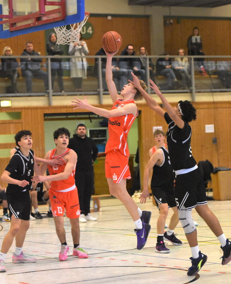 Ein junger Basketballspieler im orangefarbenen Trikot springt dynamisch zum Korb während ein Gegner in Schwarz versucht den Wurf zu blocken