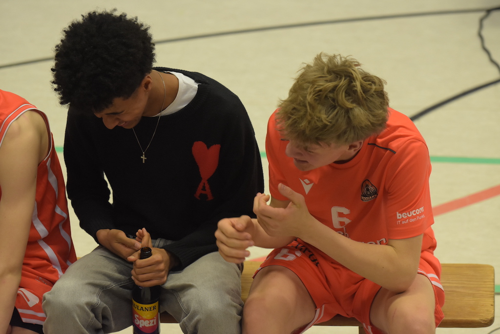 Zwei Jugendliche sitzen auf einer Bank, einer trägt Schwarz mit rotem Herz, einer Orange