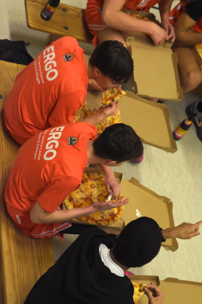 Drei junge Männer in roten Trikots essen lachend Pizza aus Kartons auf einer Holzbank