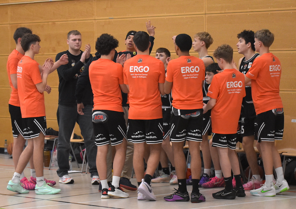 Eine Gruppe junger Basketballspieler in orangefarbenen Trikots feiert im Kreis im Sporthallenlicht