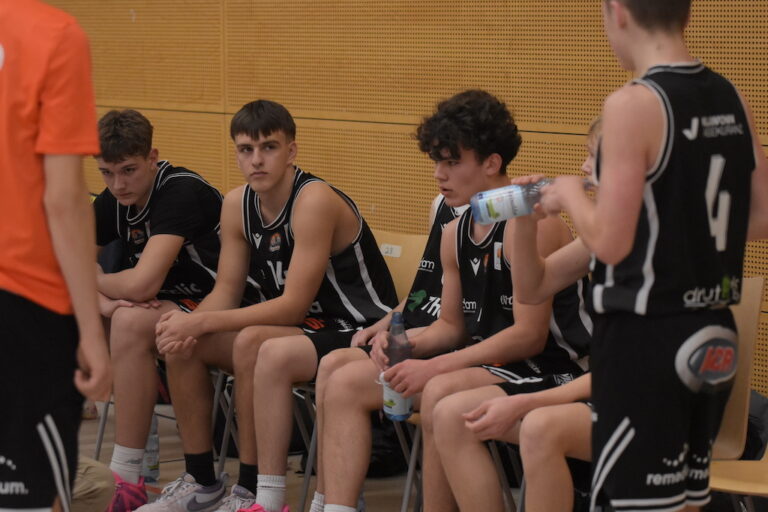 Vier junge Basketballspieler in schwarzen Trikots sitzen konzentriert auf der Bank