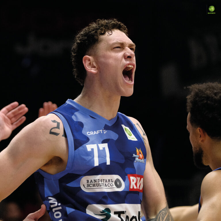 Ein junger Basketballspieler mit lockigem Haar und tätowiertem Arm jubelt laut