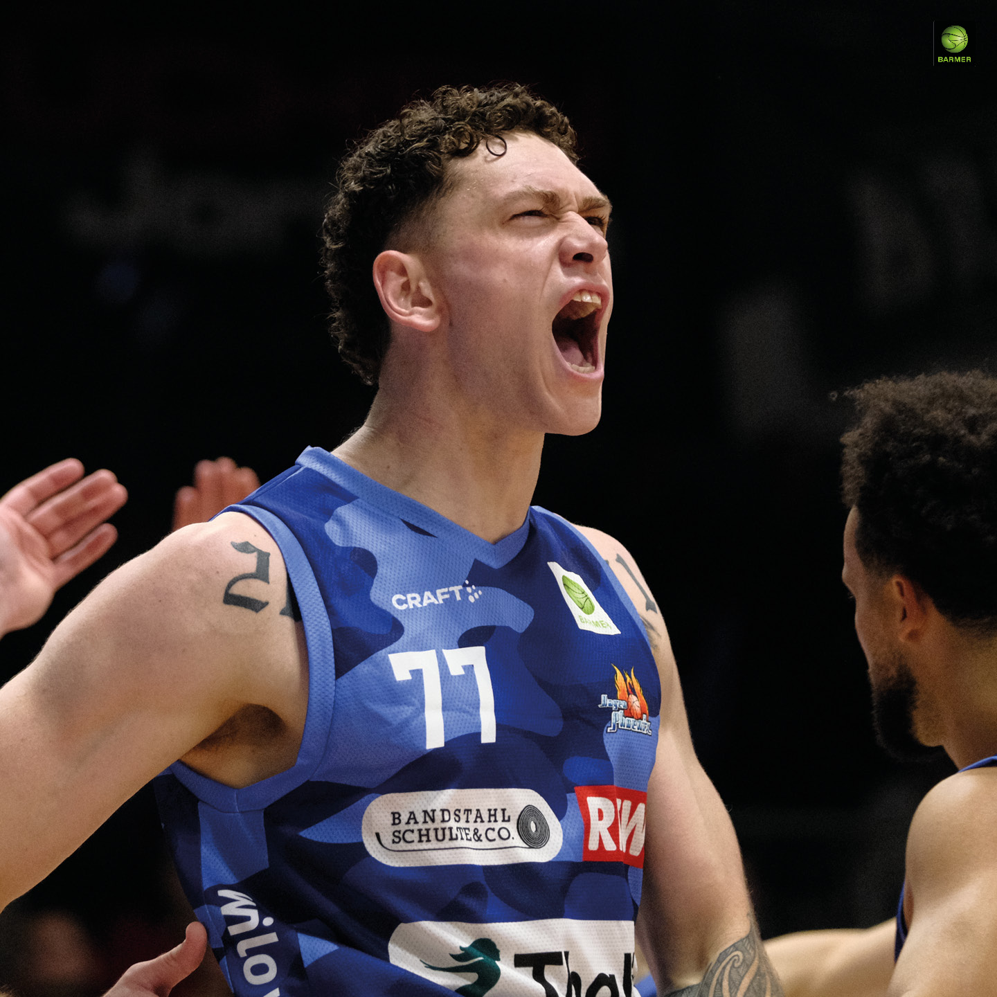 Ein junger Basketballspieler mit lockigem Haar und tätowiertem Arm jubelt laut