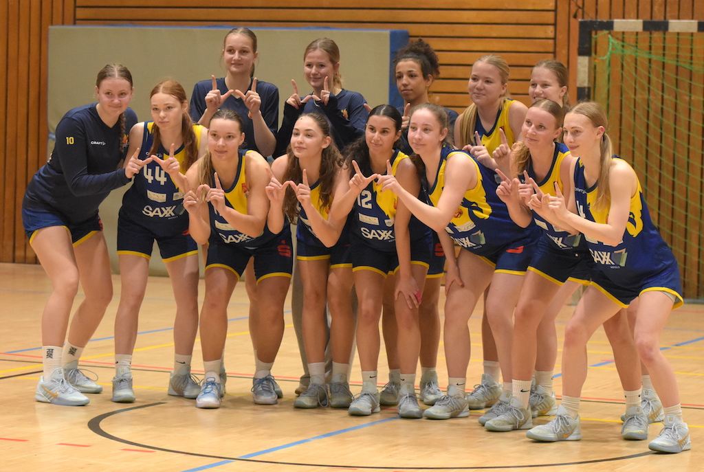 Eine Basketball Damenmannschaft in blauen und gelben Trikots posiert lachend in der Halle