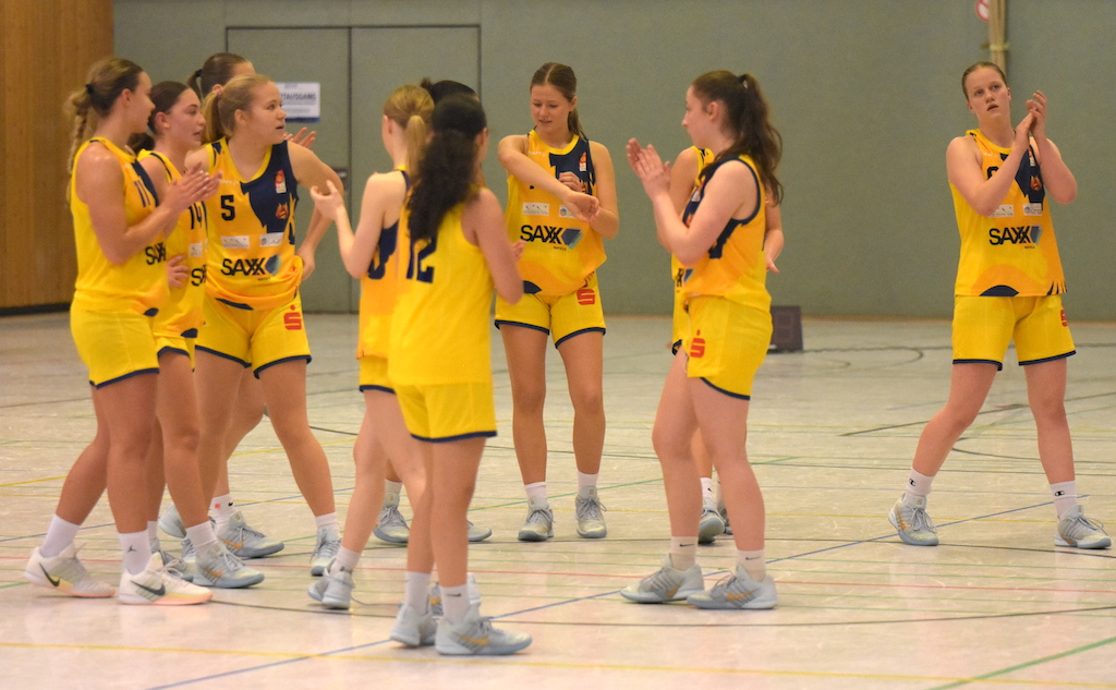 Eine Frauen Basketballmannschaft in gelben Trikots steht lachend im Kreis und klatscht