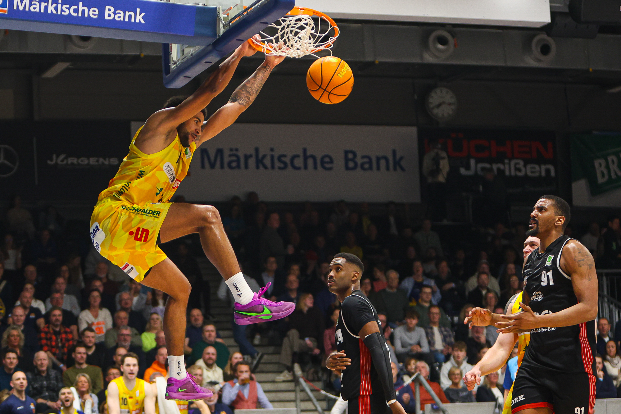 Ein Basketballspieler im gelben Trikot dunkt kraftvoll, Publikum schaut gespannt