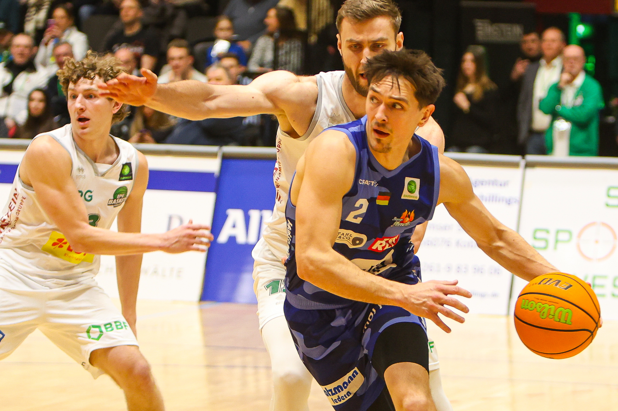 Ein Basketballspieler im blauen Trikot dribbelt konzentriert, während zwei Gegner in Weiß verteidigen