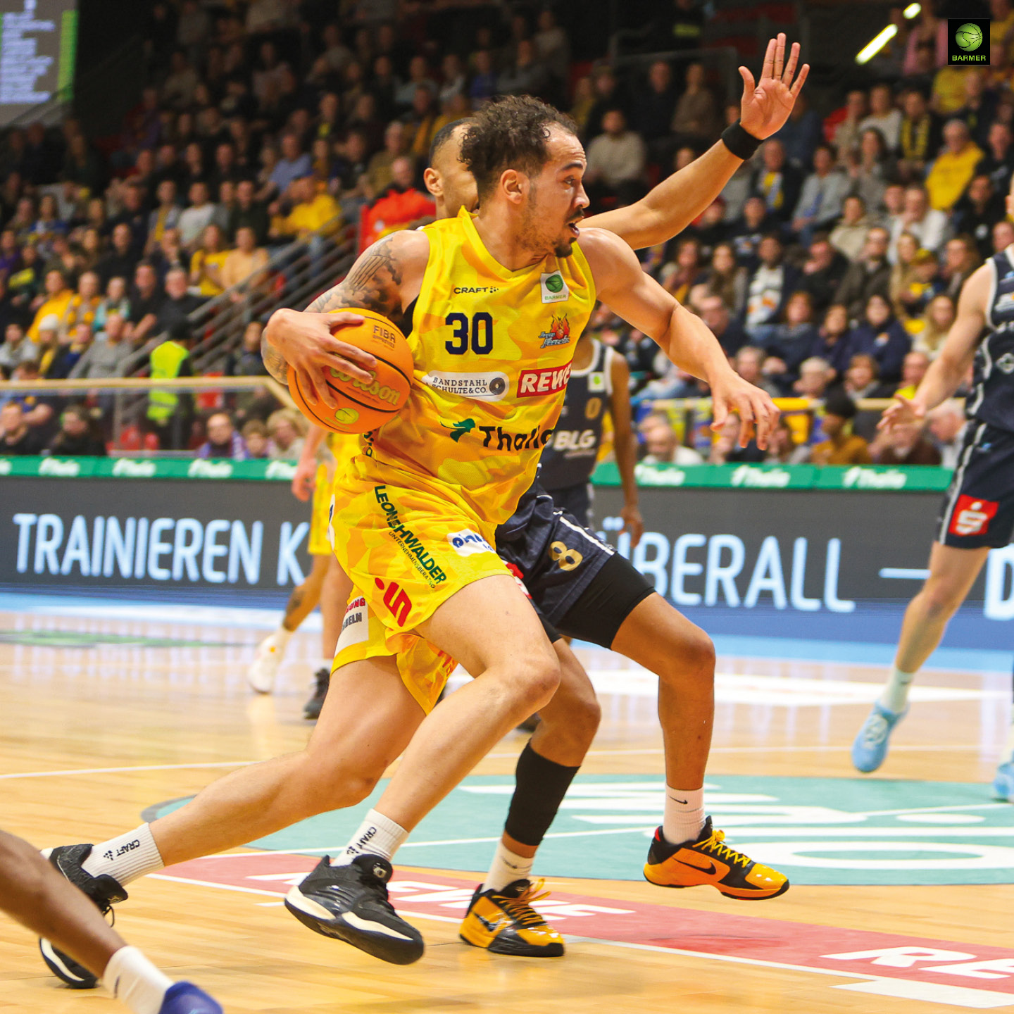 Ein Basketballspieler im gelben Trikot dribbelt konzentriert vor vollen Zuschauerrängen