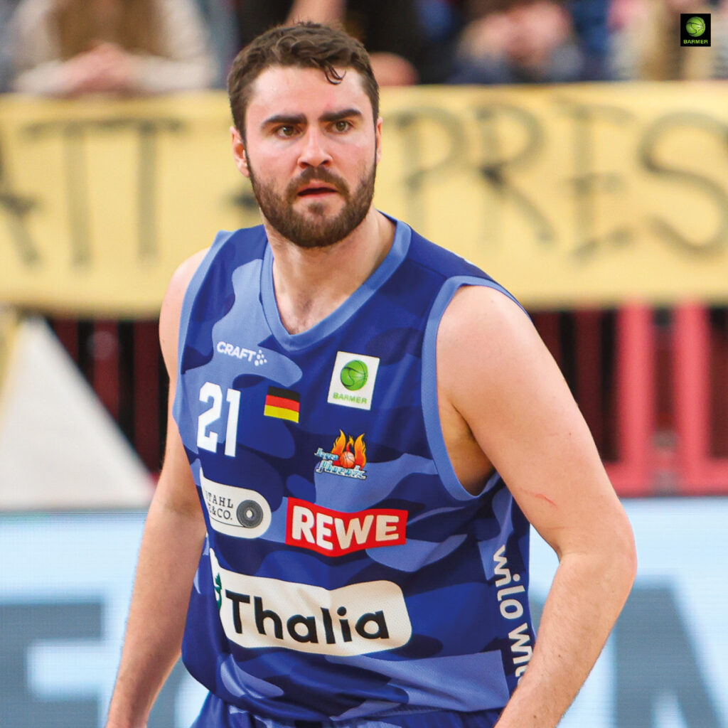 Ein Basketballspieler mit Bart und kurzem Haar trägt ein blaues Trikot wirkt fokussiert