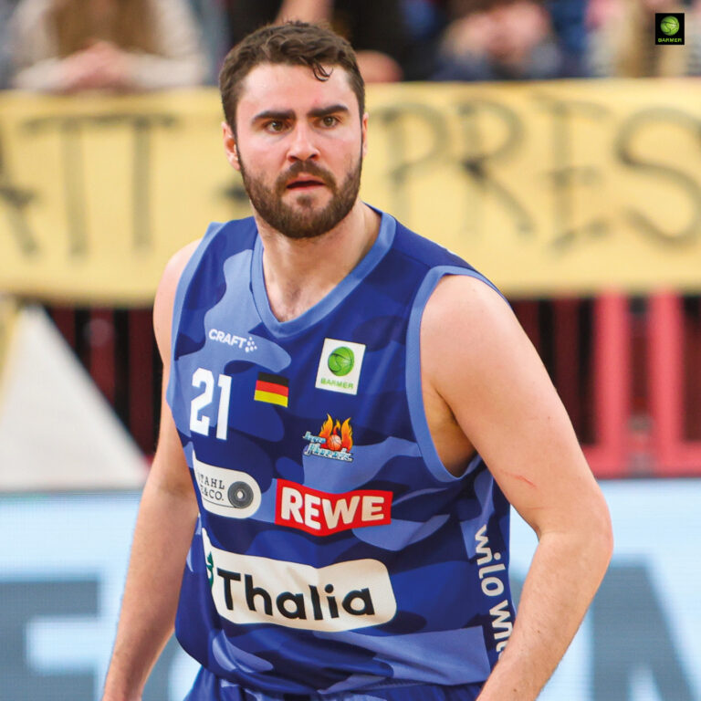 Ein Basketballspieler mit Bart und kurzem Haar trägt ein blaues Trikot wirkt fokussiert