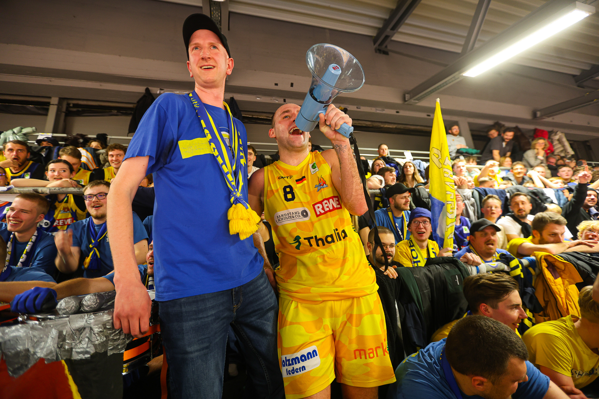 Ein lächelnder Basketballspieler im gelben Trikot jubelt mit einem Megafon im vollen Fanblock