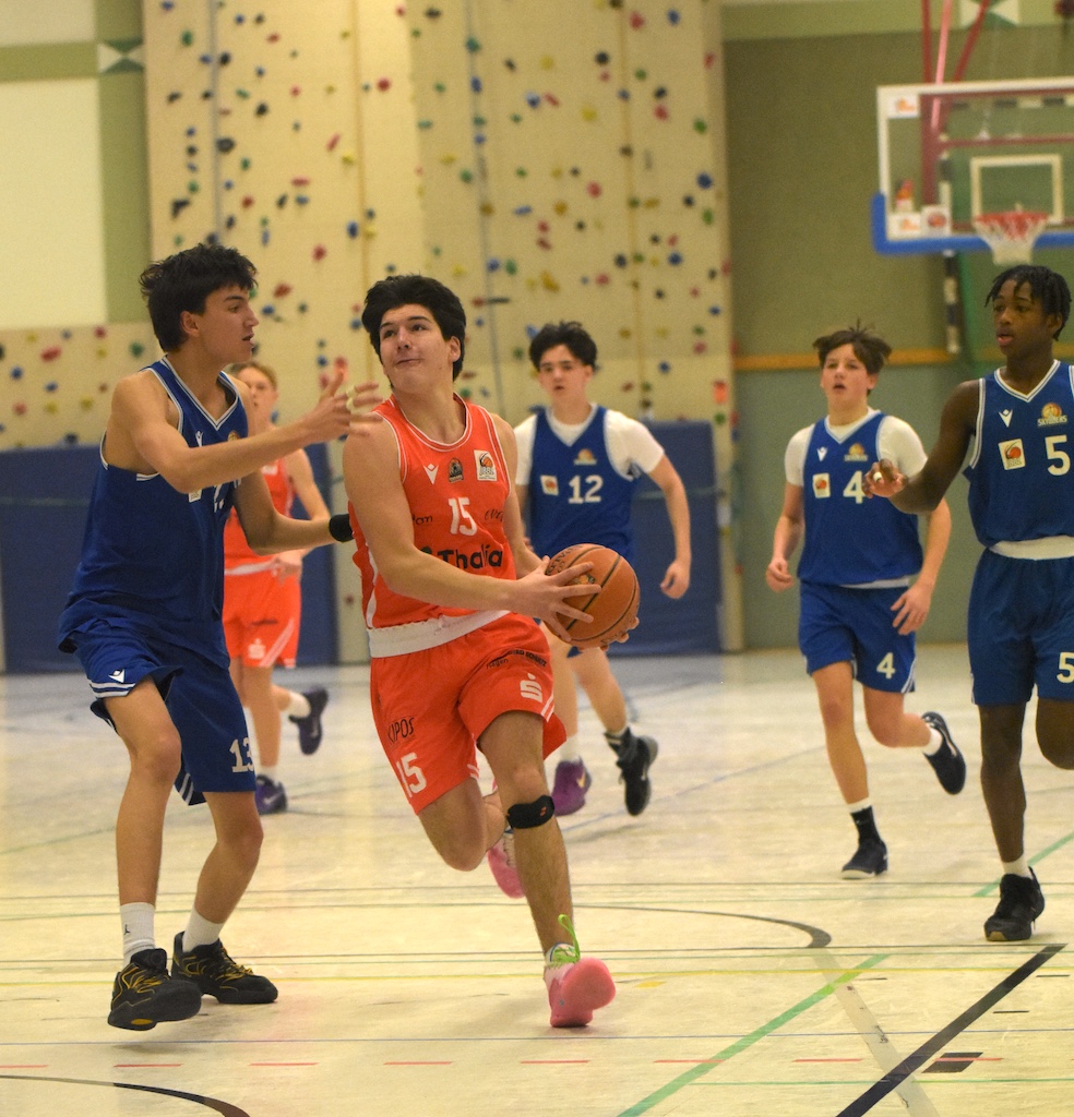 Ein junger Mann im orangefarbenen Trikot läuft konzentriert mit dem Basketball Richtung Korb, während mehrere Spieler in blauen Trikots ihn verteidigen