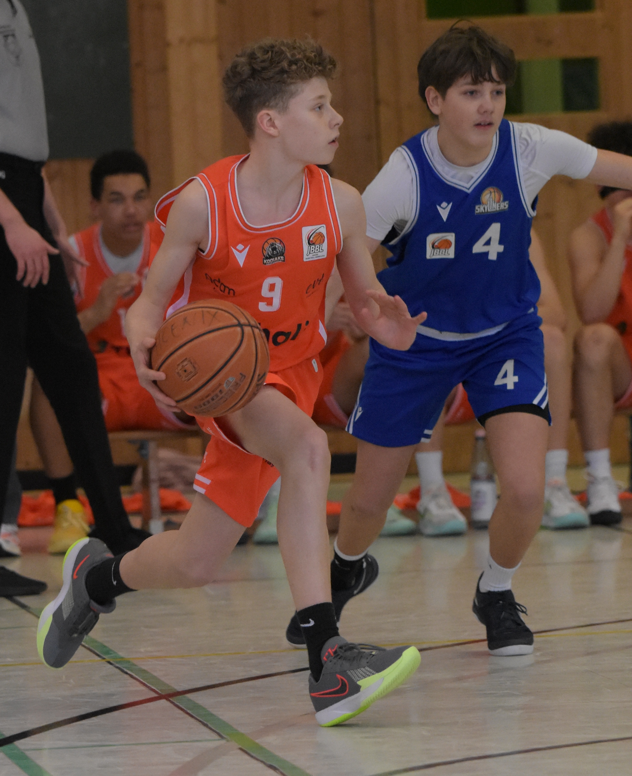 Ein Junge im orangefarbenen Basketballtrikot hält konzentriert den Ball und wird von einem Gegenspieler in blauem Trikot verteidigt