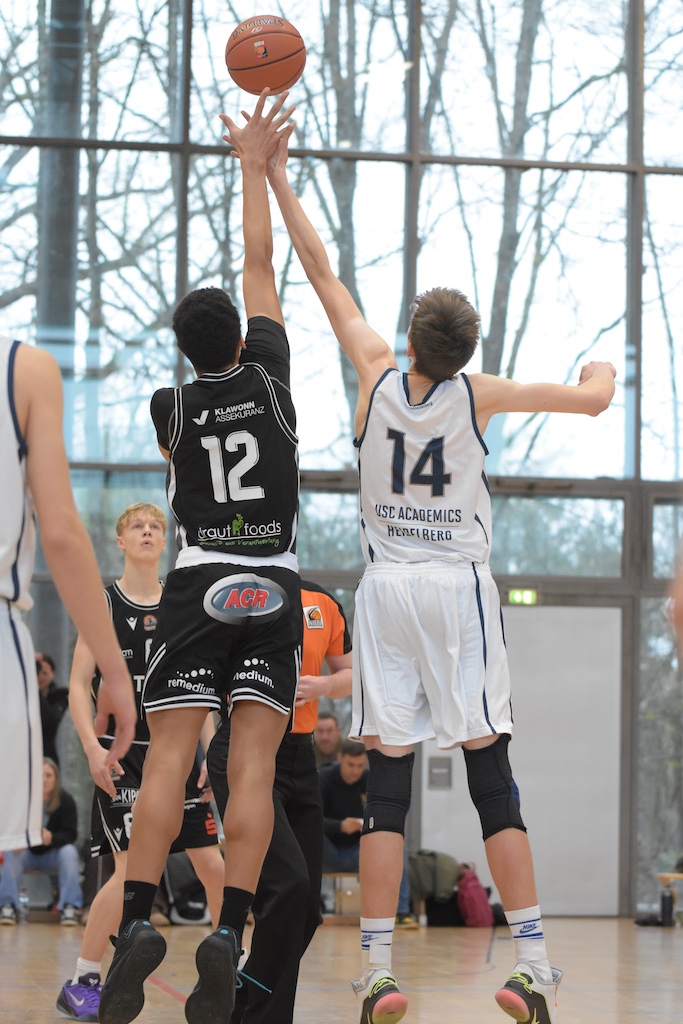 Zwei junge Basketballspieler, einer in Schwarz und einer in Weiß, springen zum Ball hoch