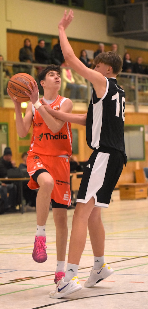 Zwei Jugendliche spielen Basketball, einer mit orangefarbenem Trikot hebt zum Korbleger ab