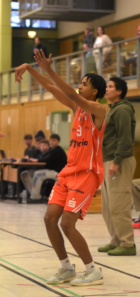 Ein junger Basketballspieler im orangefarbenen Trikot wirft konzentriert den Ball