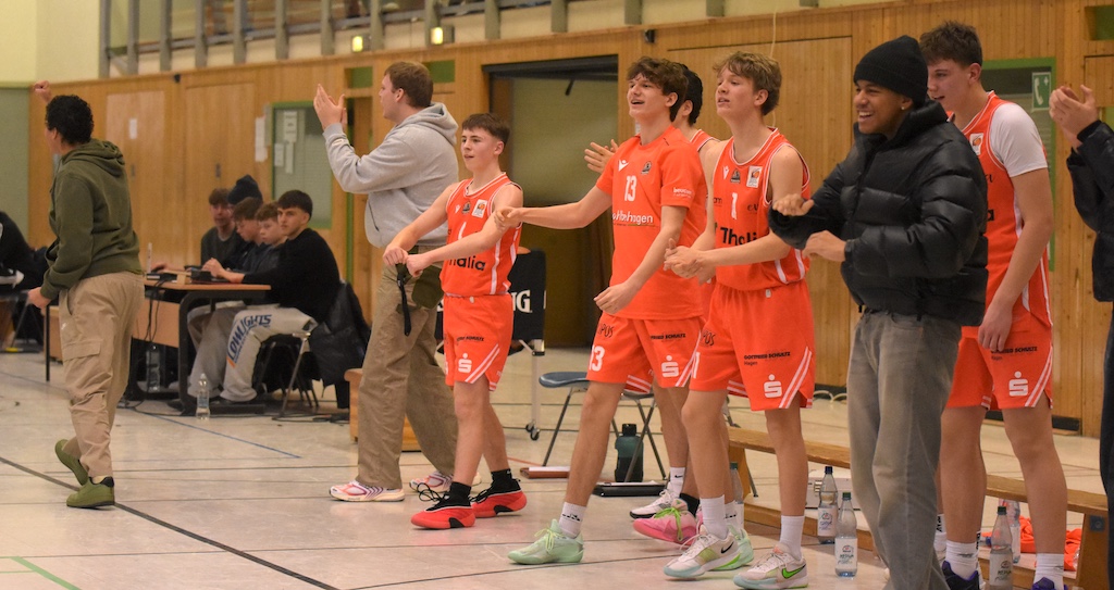 Junge Basketballspieler im orangefarbenen Trikot jubeln lachend auf der Bank