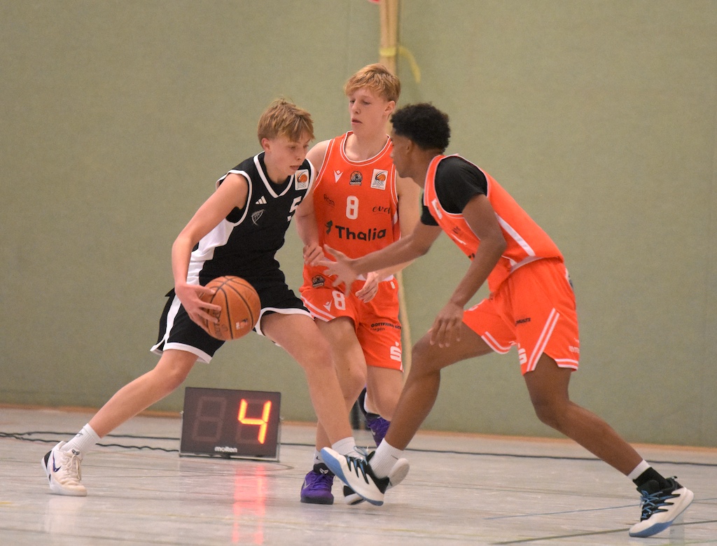Drei junge Basketballspieler, zwei in Orange und einer in Schwarz, kämpfen konzentriert.