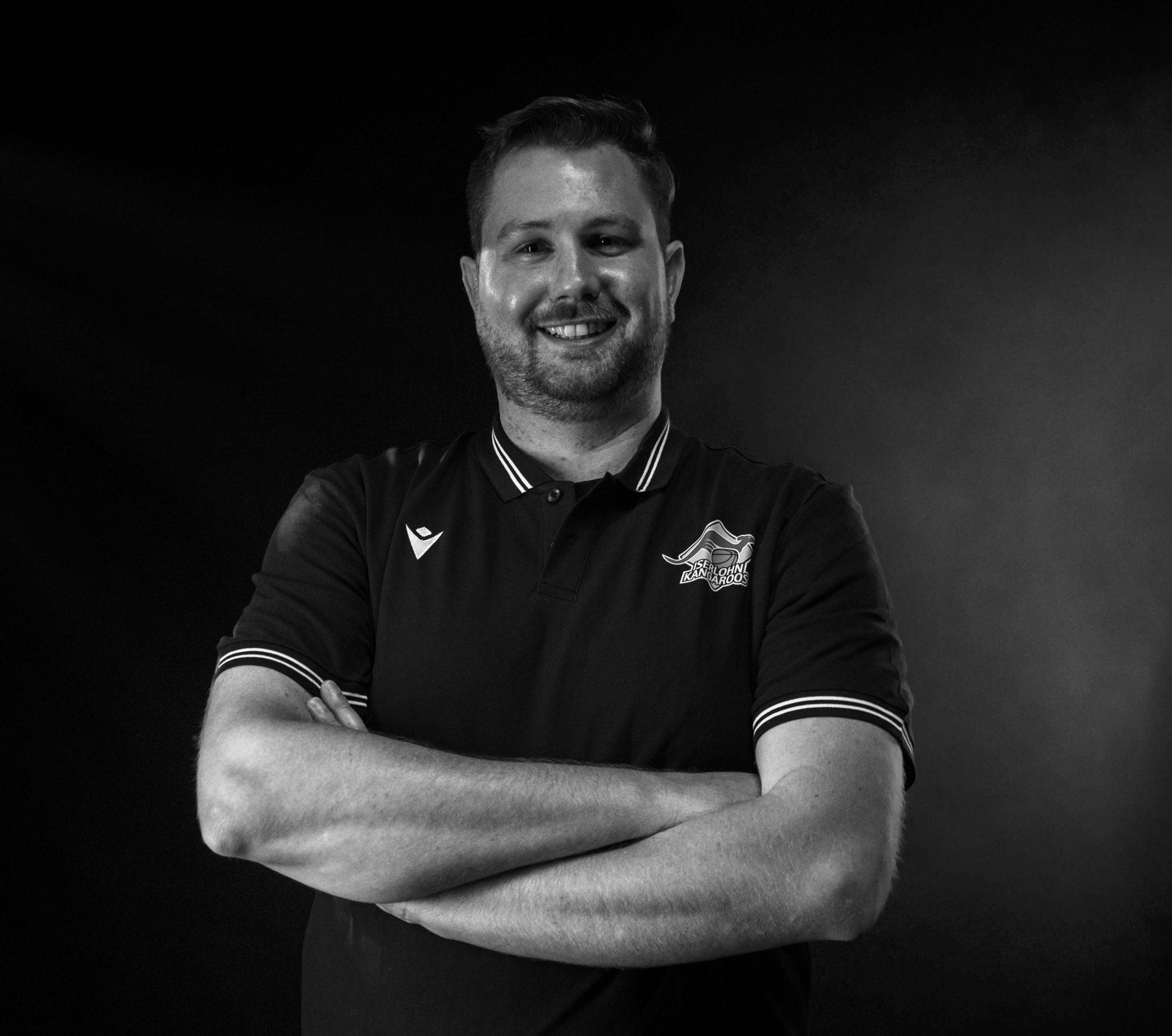 Ein lächelnder Mann mit Bart trägt ein dunkles Poloshirt und verschränkt die Arme