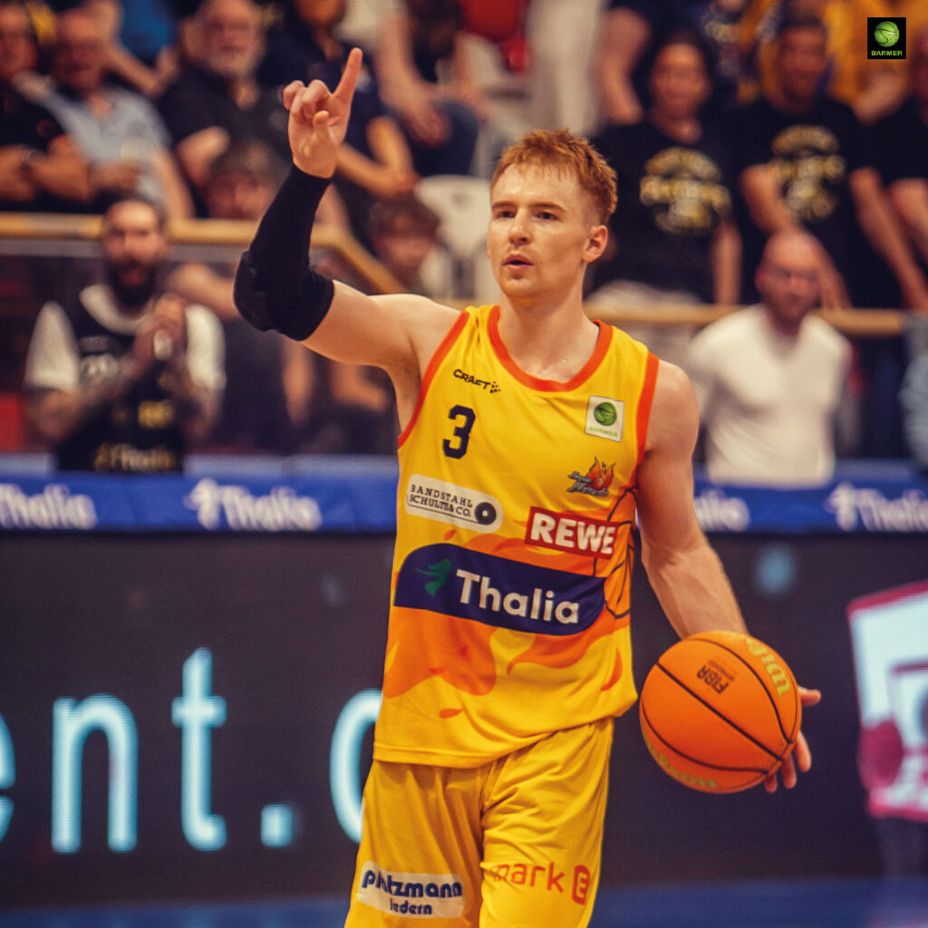 Ein junger Basketballspieler im gelben Trikot hebt ernst den Arm und hält den Ball