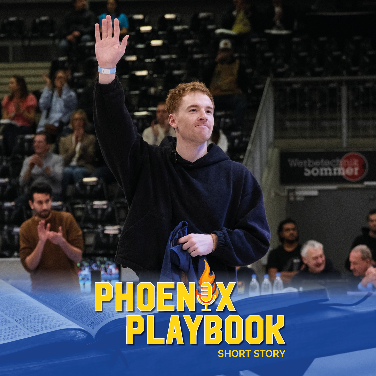 Junger Mann mit blassem Teint und rötlichen Haaren winkt freundlich in dunkler Halle