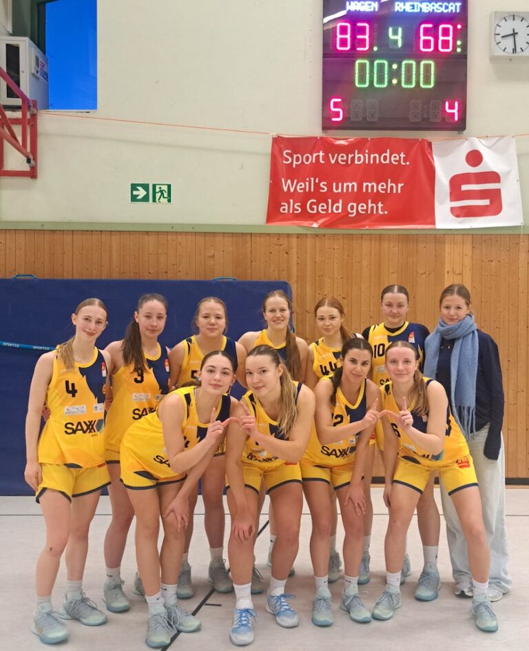 Eine Basketballspielerin mit blondem Zopf und ernster Miene steht mit ihrem Team im gelben Trikot vor einer Anzeigetafel, die ein gewonnenes Spiel zeigt