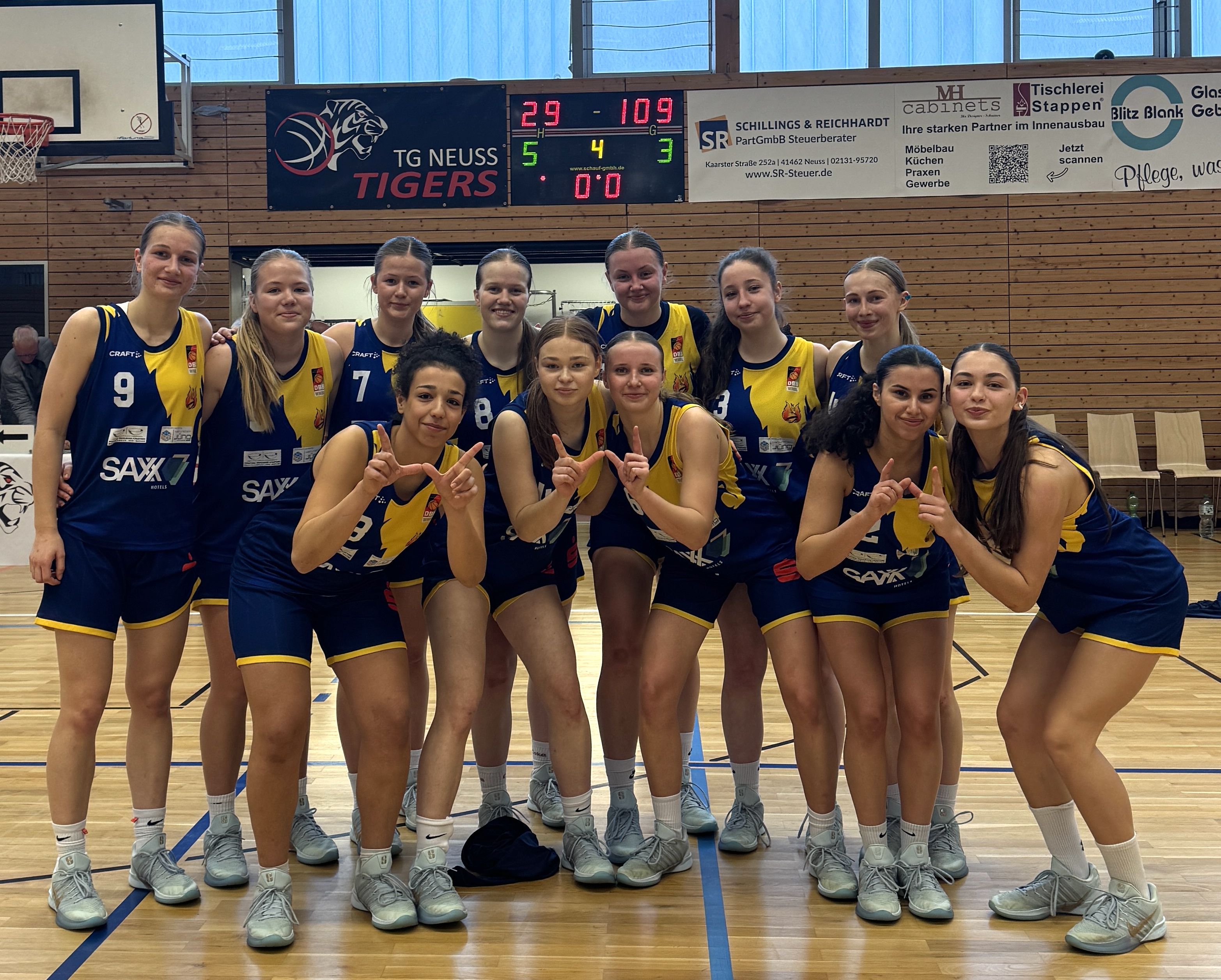 Elf junge Frauen im blauen Basketballtrikot lächeln siegreich und stehen Arm in Arm auf dem Hallenboden