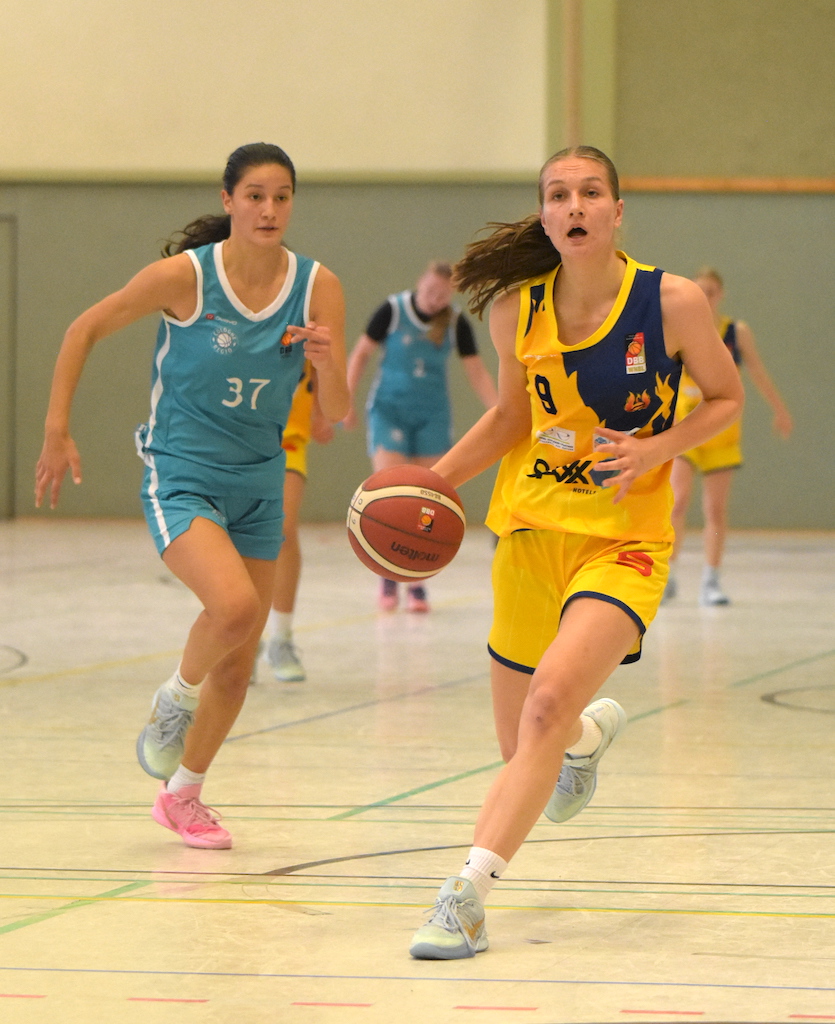 Eine junge Frau im gelben Basketballtrikot läuft entschlossen mit dem Ball übers Feld