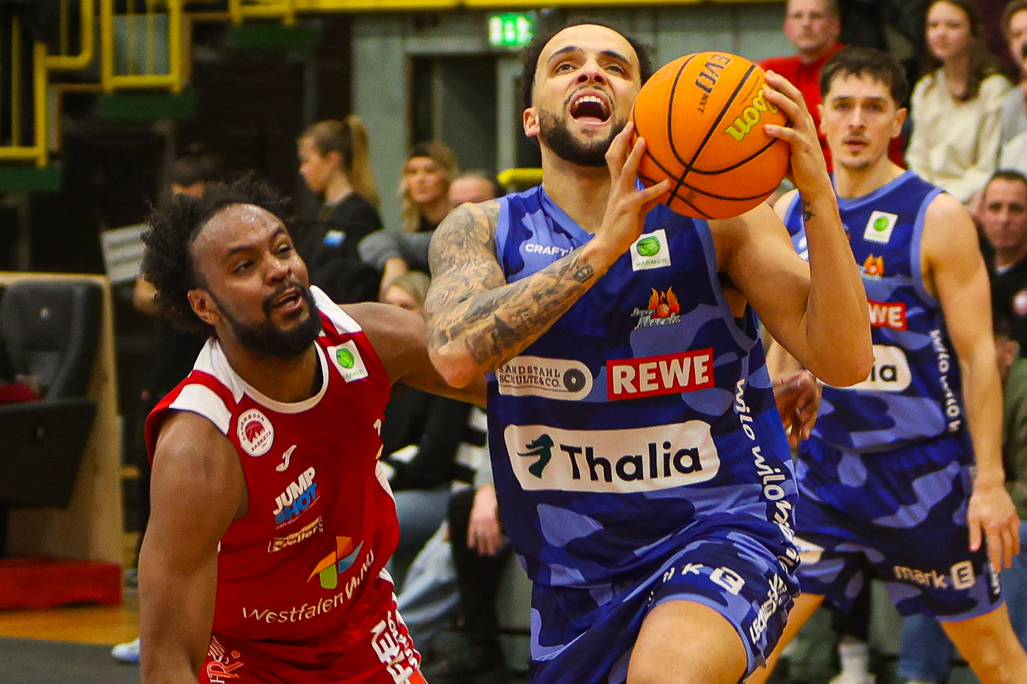 Ein Basketballspieler im blauen Trikot geht entschlossen zum Korb, verfolgt von einem Gegner in Rot