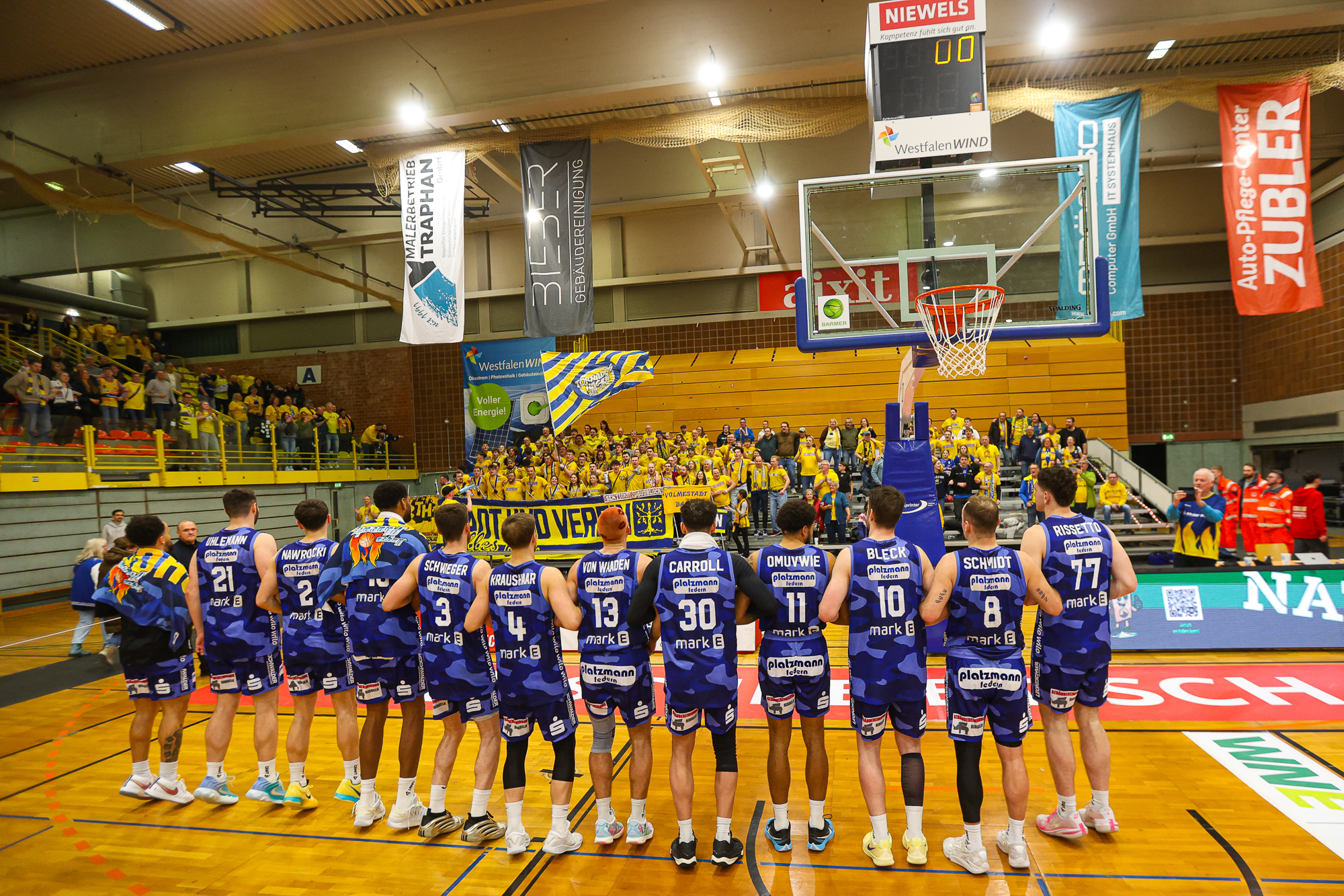 Das Herrenhandballteam in blauen Trikots steht lächelnd Arm in Arm vor gelben Fans