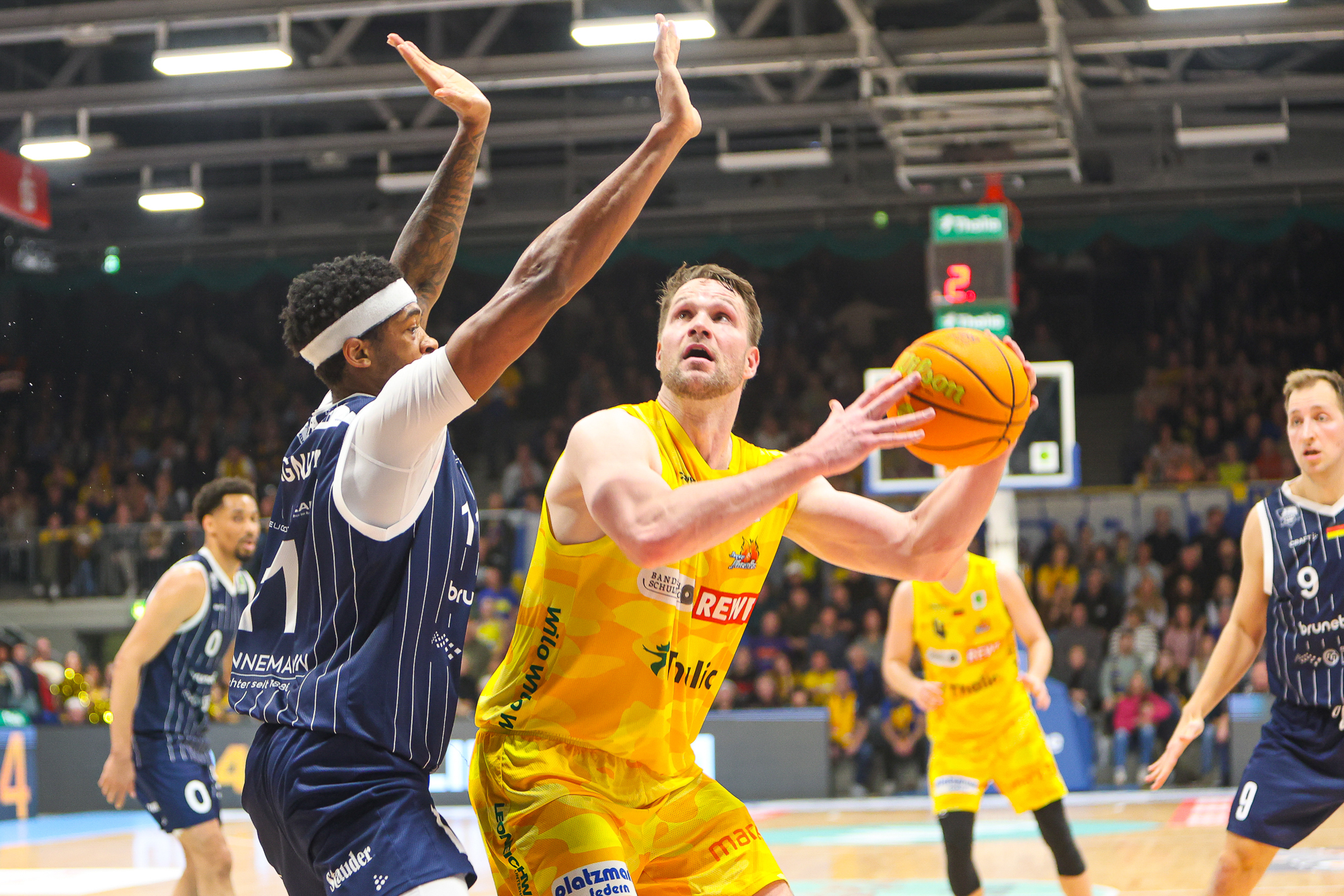 Ein Basketballspieler im gelben Trikot schaut fokussiert zum Korb und wird von einem Gegenspieler in blau mit nach oben gestreckten Armen verteidigt