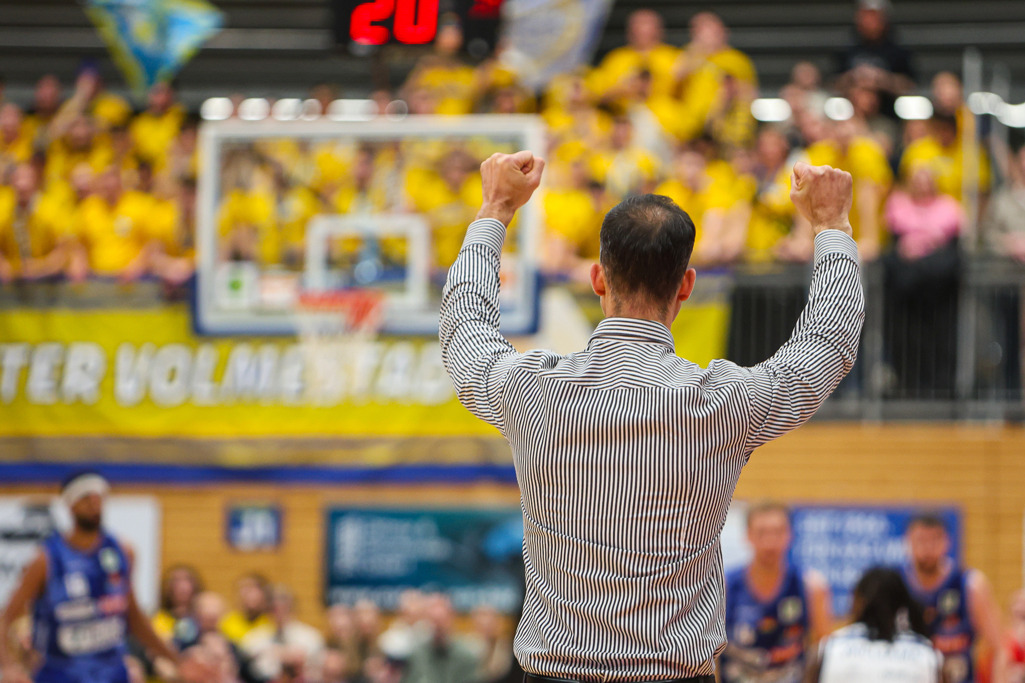 Ein Mann mit gestreiftem Hemd hebt jubelnd die Arme vor einer Menge in Gelb