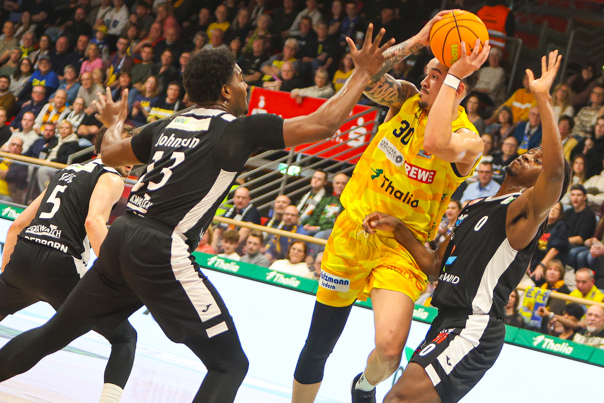 Zwei Basketballspieler in schwarzen Trikots verteidigen einen Gelbgekleideten beim Wurf