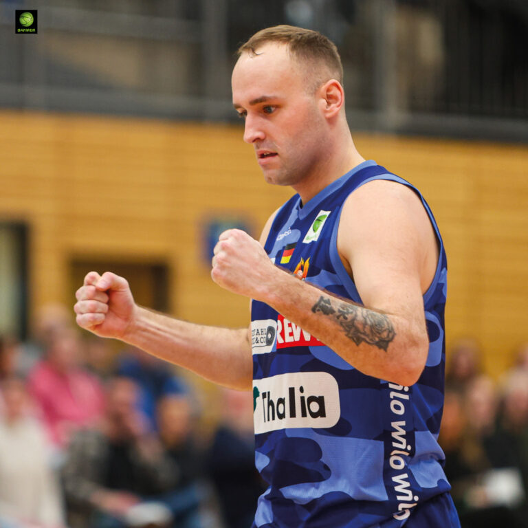 Ein konzentrierter Basketballspieler im blauen Trikot ballt die Fäuste und zeigt ein Tattoo