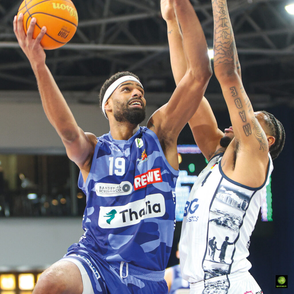 Ein Basketballspieler im blauen Trikot springt entschlossen zum Korb, verteidigt von einem Spieler in Weiß