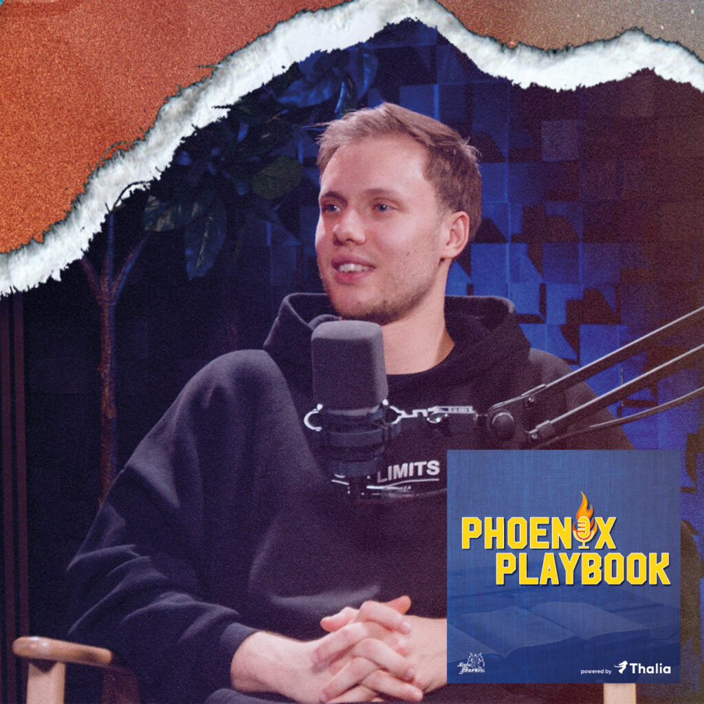 Ein junger Mann mit blondem Haar und freundlichem Gesichtsausdruck sitzt vor einem Mikrofon und trägt einen schwarzen Hoodie