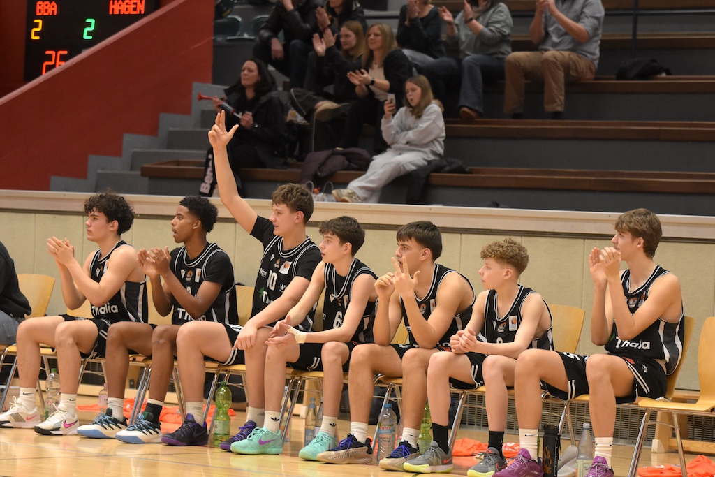 Sieben junge Basketballspieler im schwarzen Trikot sitzen auf der Bank und jubeln lebhaft