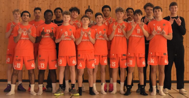 Vierzehn junge Männer in orangefarbenen Trikots posieren lächelnd vor einer Holzvertäfelung