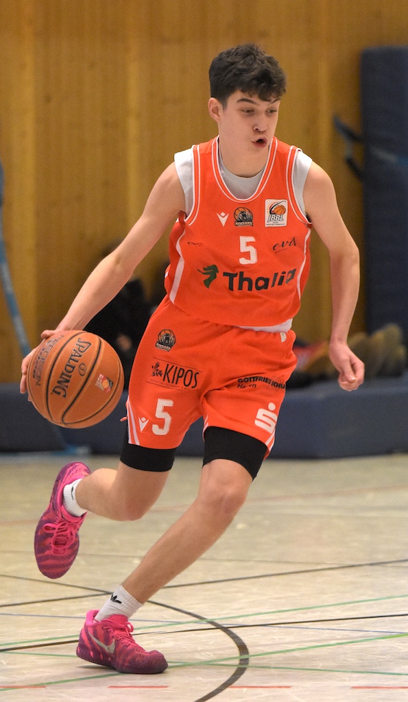 Ein junger Basketballspieler im orangefarbenen Trikot mit konzentriertem Gesichtsausdruck dribbelt den Ball auf einem Hallenboden