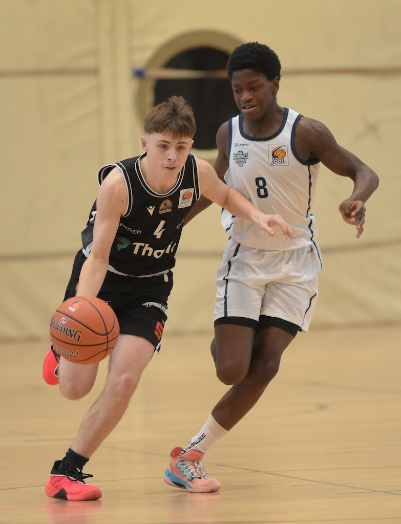 Zwei junge Basketballspieler im Spiel, einer konzentriert dribbelnd, der andere verteidigt energisch, beide in Sporttrikots, das Spielfeld ist hell und gut beleuchtet