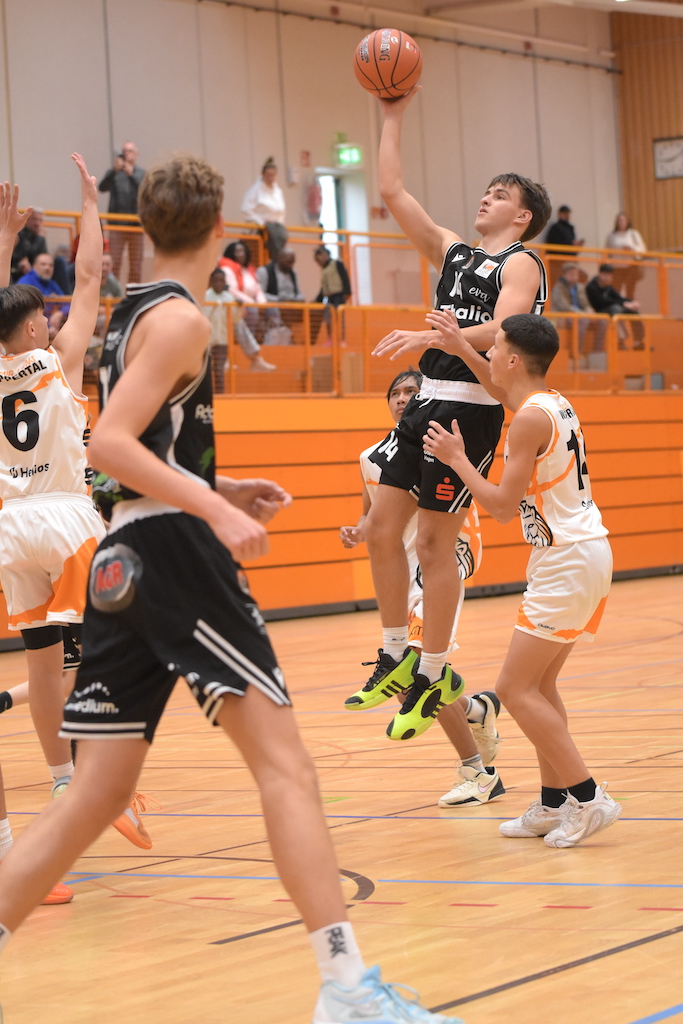 Ein junger Basketballspieler im schwarzen Trikot springt konzentriert zum Korbleger, umgeben von Gegenspielern in weißen Trikots auf einem Hallenparkett